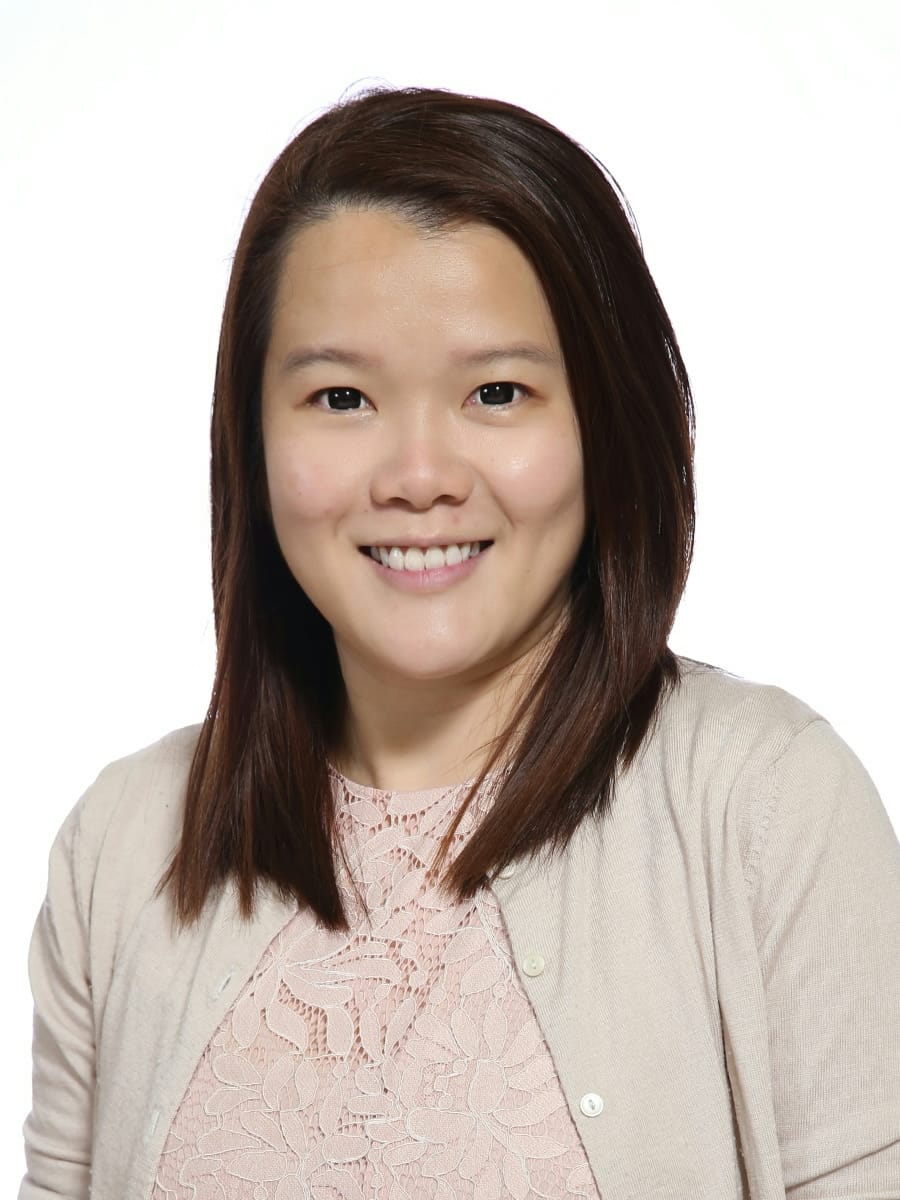 A smiling woman with straight dark brown hair wearing a pink lace top and beige cardigan against a white background.
